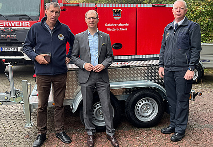 Drei Männer stehen vor einem kleinen zweiachsigen Anhänger.