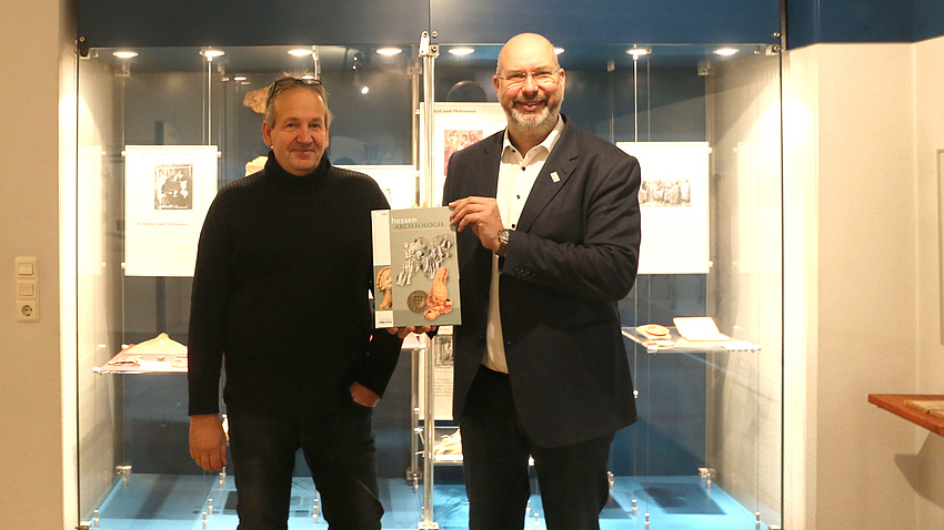 Zwei Männer stehen in einem Museum. Hinter ihnen eine große mannshohe Vitrine mit Ausstellungsstücken. Einer der Männer hält ein Buch in der Hand.