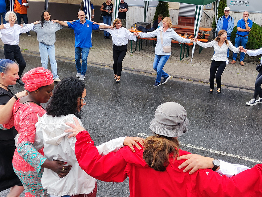 Menschen unterschiedlichen Alters, Geschlechts, Hautfarbe und Kultur tanzen im Kreis auf der Straße. Dabei halten sie sich an den Schultern 