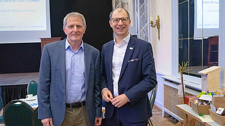 Zwei Männer stehen in einem Saal nebeneinander.