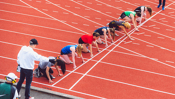 Auf der Tartanbahn eines Sportplatzes stehen mehrere Athletinnen in den Startblöcken.