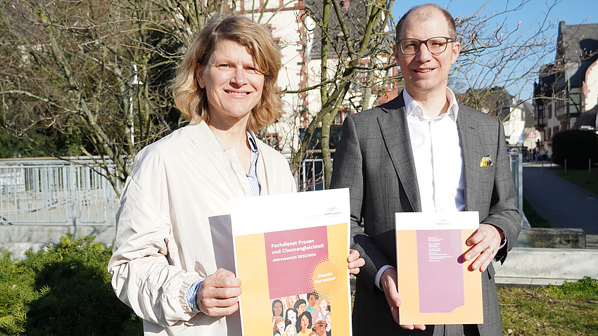 Eine Frau und ein Mann stehen auf einer Wiese vor einem Metallgeländer. Im Hintergrund sind Häuser zu sehen. Die Frau hält ein Plakat in der Hand, der Mann eine Broschüre.