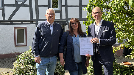 Der Landrat übergibt den Bescheid der Familie, im Hintergrund ist das Haus zu sehen.