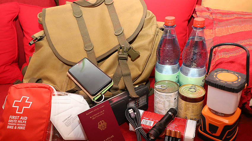 Das Bild zeigt einen Rucksack, haltbare Lebensmittel, Medikamente und die wichtigsten persönlichen Dokumente wie den Reisepass.