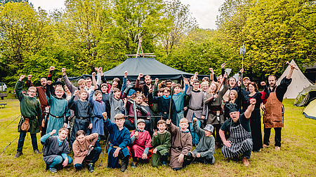 Eine Personengruppe in mittelalterlicher Gewandung. Die meisten stehen, einige in der Hocke.