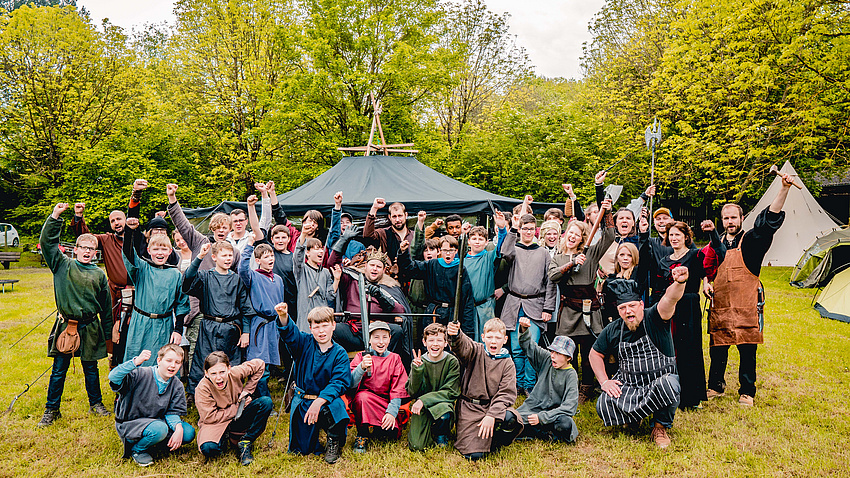 Eine Personengruppe in mittelalterlicher Gewandung. Die meisten stehen, einige in der Hocke.