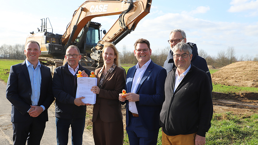 Ein Frau und fünf Männer stehen gemeinsam auf einem Feld. Im Hintergrund ist ein Bagger zu sehen. Die Frau und einer der Männer halten gemeinsam ein dickes Paket Akten in der Hand.