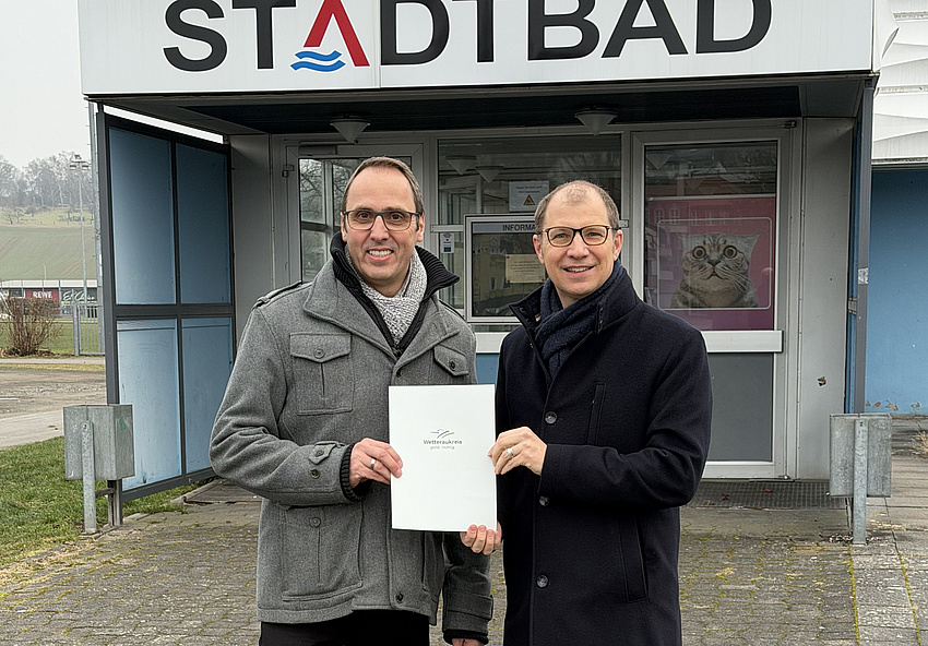 Zwei Männer stehen vor dem Eingang eines Gebäudes, an dem ein Schild mit dem Begriff Stadtbad angebracht ist. Beide halten gemeinsam ein Dokument in Händen.