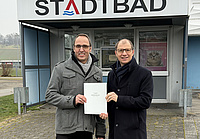 Zwei Männer stehen vor dem Eingang eines Gebäudes, an dem ein Schild mit dem Begriff Stadtbad angebracht ist. Beide halten gemeinsam ein Dokument in Händen.