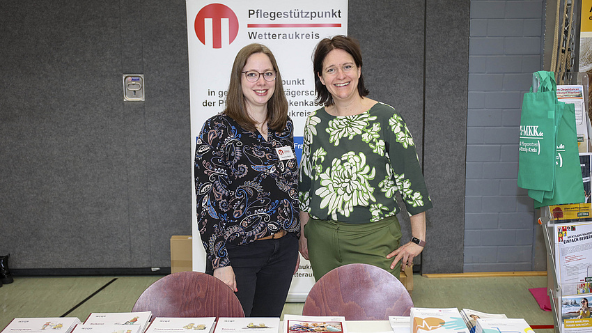 Zwei Frau stehen hinter einem Infostand, auf dem viele Broschüren ausgelegt sind. Hinter ihnen steht ein Banner, auf dem "Pflegestützpunkt Wetteraukreis" zu lesen ist.