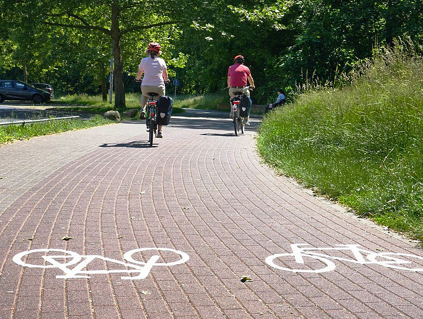 Blick auf zwei Radfahrer, die unterwegs sind auf einem städtischen Radweg, der am Grunde mit Radsymbolen markiert ist.