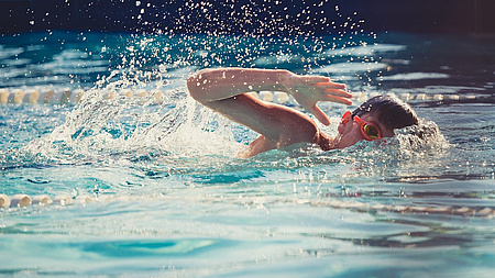 Ein Junge schwimmt im Wasser. Er krault undträgt eine Schwimmbrille.