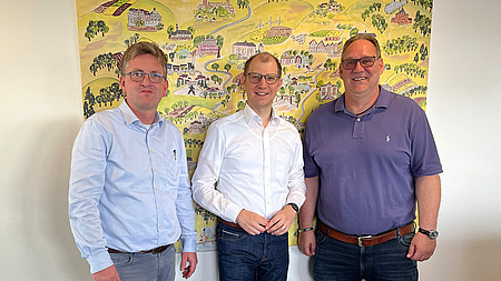 Drei Männer stehen nebeneinander und lächeln in die Kamera. Im Hintergrund ein in naiver Kunst gemaltes Bild.