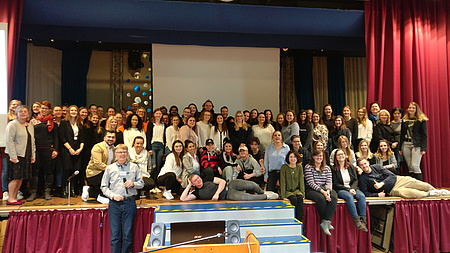 Eine große Gruppe junger Menschen steht und sitzt auf einer Bühne. In der Mitte davor führt eine kleine Treppe auf die Bühne.