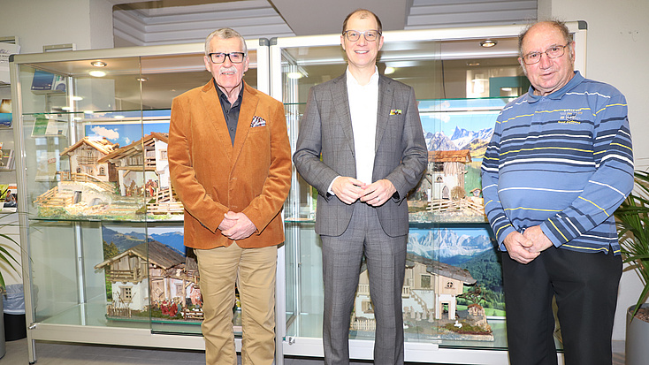 Drei Männer stehen in einem größeren Raum vor einer Vitrine, in der mehrere Weihnachtskrippen zu sehen sind.