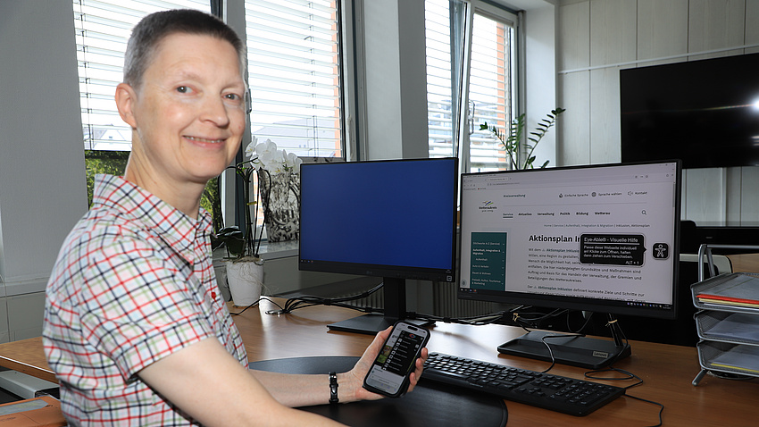 Eine Frau sitzt in einem Büro an einem Schreibtisch. Auf diesem stehen vor ihr zwei Computer-Bildschirme und eine Tastatur. In der Hand hält sie ein Smartphone.