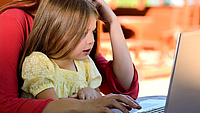 Kleines Mädchen auf dem Schoß einer Frau vor einem aufgeklappten Laptop