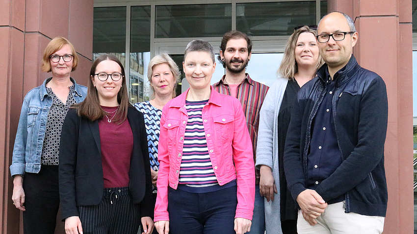 Sieben Personen, fünf Frauen und zwei Männer, stehen lächelnd vor dem Eingang eines Gebäudes.