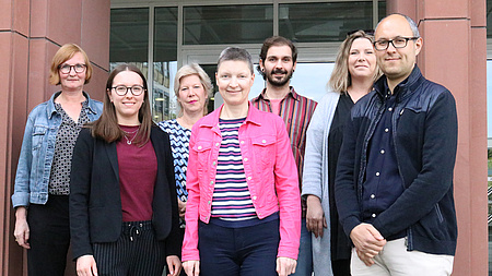 Sieben Personen, fünf Frauen und zwei Männer, stehen lächelnd vor dem Eingang eines Gebäudes.