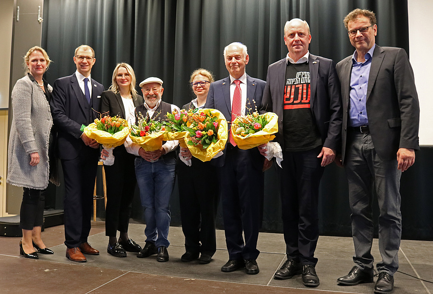 Mehrere Männer und Frauen stehen nebeneinander auf einer Bühne. Fünf Personen von ihnen haben einen Blumenstrauß in der Hand.