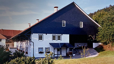 Das Hauptgebäude eines Bauernhofs steht am Rande einer Wiese und eines Waldes.
