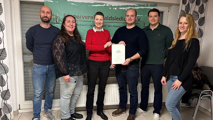 Drei Frauen und drei Männer stehen in einem Raum. Eine der Frauen und einer der Männer halten gemeinsam ein Dokument in den Händen.