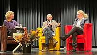 Zwei Frauen und ein Mann auf einer Bühne. Jede Person sitzt auf einem andersfarbigen Sessel. Der Mann in der Mitte spricht in ein Mikrofon.