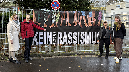 Vier Frauen stehen vor einem Plakat mit der Aufschrift "Kein Platz für Rassismus".