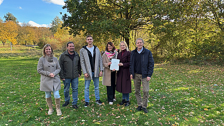 Sechs Personen, drei Frauen und drei Männer, stehen auf einem Wiesengelände. Im Hintergrund sind mehrere Bäume zu sehen. Zwei der Frauen halten ein Dokument in Händen.