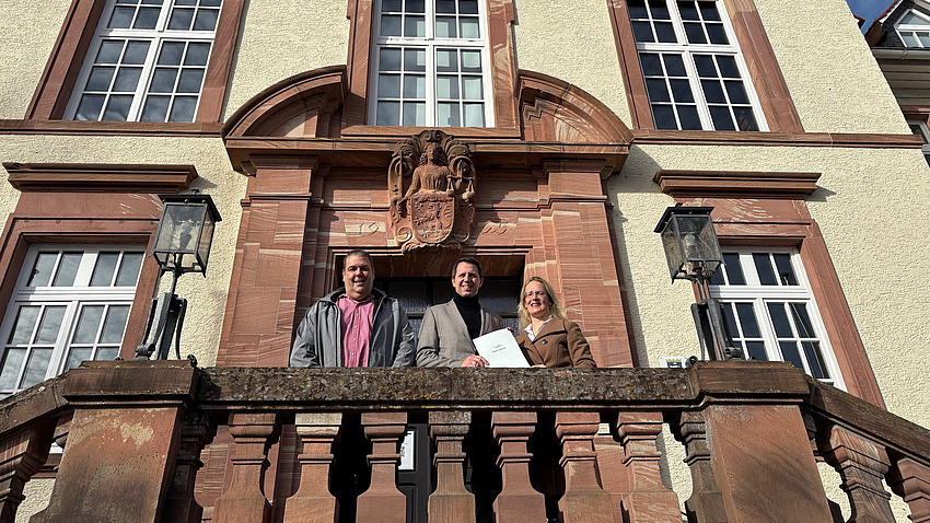 Drei Personen stehen auf einem Balkon, vor der Eingangstür in das Rathaus der gemeinde Altenstadt. Das Bild ist von unten nach oben fotografiert, sodass auch die Fassade des historisches Gebäudes erkennbar ist.