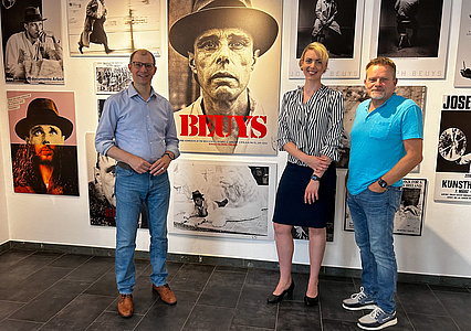 Eine Frau und zwei Männer stehen vor einer Wand mit mehreren Ausstellungsplakaten.