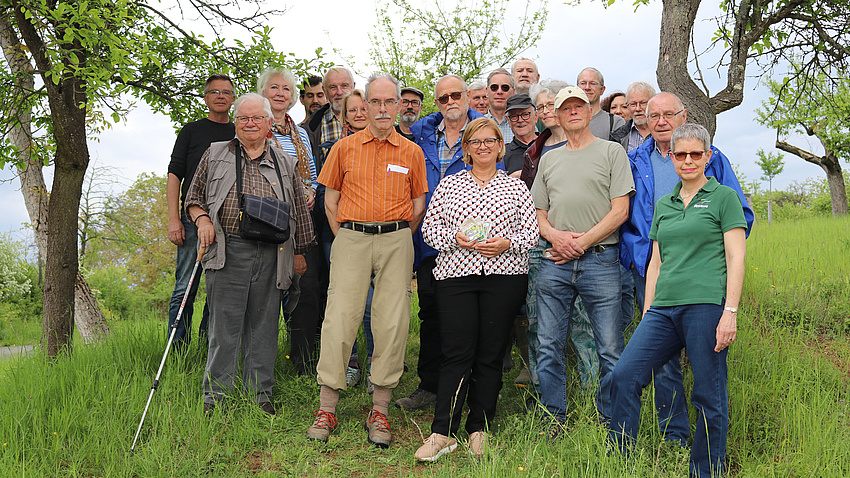 Eine Gruppe von rund 15 Männern und Frauen steht auf einem grünen Hügel. Im Hintergrund sind Bäume und Pflanzen zu sehen.