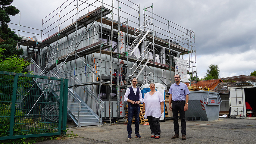 Drei Personen stehen vor einem Gebäude, um das ein Gerüst aufgebaut ist.