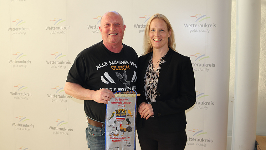 Ein Mann und eine Frau stehen vor einer Stellwand. Der Mann hält einen Wimpel in der Hand.