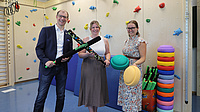 Zwei Frauen und ein Mann stehen in einer Sporthalle und halten Sportausrüstung in den Händen.