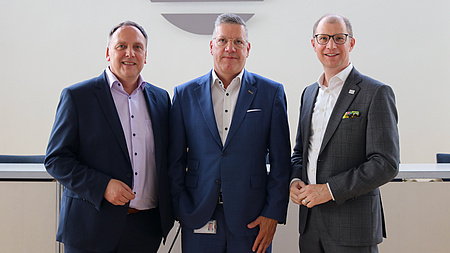 Drei Männer stehen vor einer weißen Wand. Sie lächeln in die Kamera.