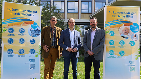 Prof. Dr. Reinhold Merbs, Landrat Jan Weckler und Dr. Alexander Jakob stehen auf einer Wiese. Neben ihnen zwei Roll-Ups zum Thema Hitzeschutz.
