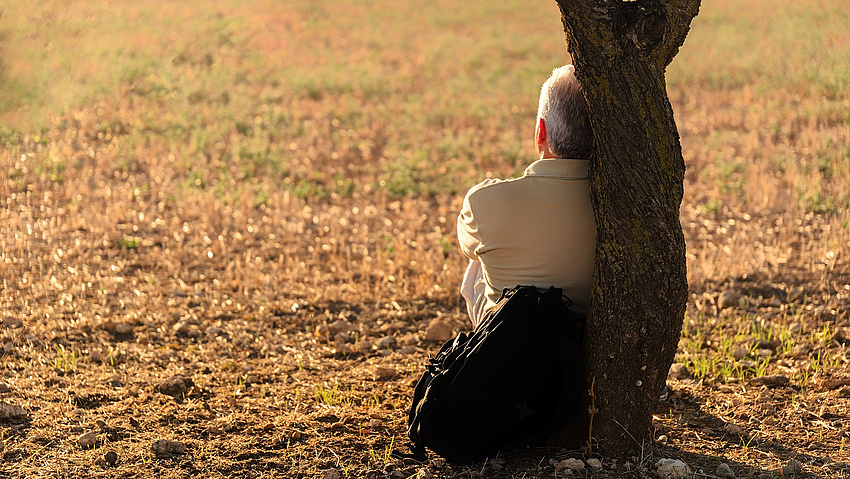 Ein alter Mann sitzt lehnt mit einem Rücken am Baum.