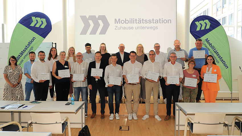 In einem großen Saal stehen mehrere Frauen und Männer in zwei Reihen vor einer Leinwand. Einige von ihnen halten Urkunden in Händen.