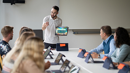 Mehrere Menschen sitzen um einen Tisch herum, einer steht vorne und erklärt etwas.