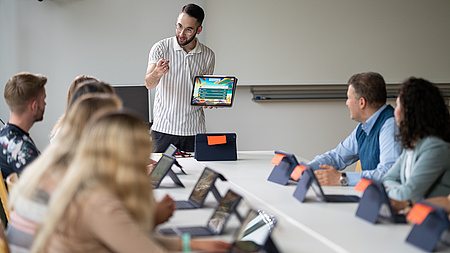Ein junger Mann steht vor einer Tischgruppe, an der andere Leute sitzen. Er erklärt etwas.