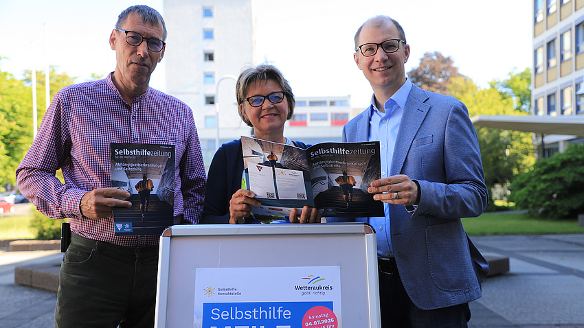 Eine Frau und zwei Männer stehen im Freien auf einem großen Platz. Vor der Gruppe steht ein Plakat und alle drei halten Zeitschriften in Händen.