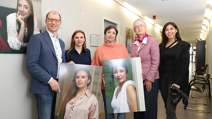 Vier Frauen und ein Mann stehen in einem Gang, an dessen einer Wand Porträtfotos von Frauen hängen. Zwei weitere Porträtfotos hält die Gruppe in Händen.