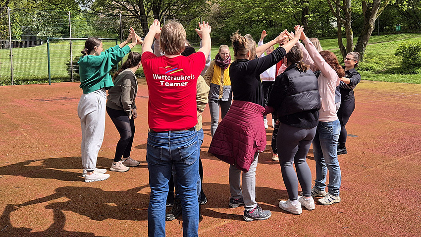 Mehrere Jugendliche stehen auf einem Sportplatz in einem Kreis.