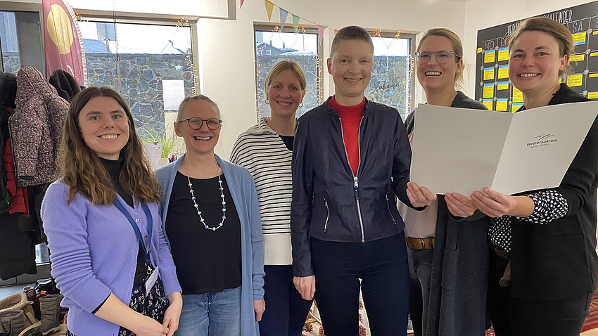 Sechs Frauen stehen nebeneinander in einem Raum und lächeln. Die drei Frauen rechts halten gemeinsam ein Dokument in den Händen.
