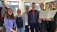 Sechs Frauen stehen nebeneinander in einem Raum und lächeln. Die drei Frauen rechts halten gemeinsam ein Dokument in den Händen.
