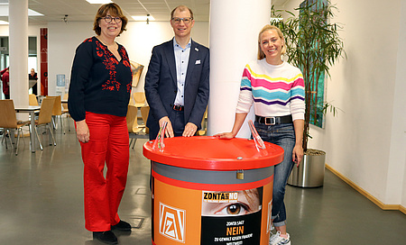 Zwei Frauen und ein Mann stehen vor einer großen Spendendose.