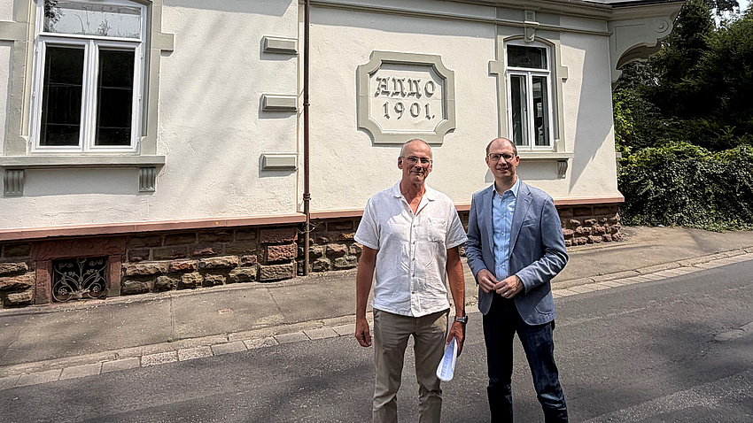 Zwei Männer stehen vor der Fassade eines alten Gebäudes, auf dem die Jahreszahl "1901" zu erkennen ist.