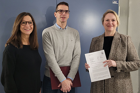 Zwei Frauen und ein Mann stehen vor einer geschlossenen Tür. Die rechts stehende Frau hält ein Schriftstück in der Hand, der Mann links daneben eine Aktenmappe.