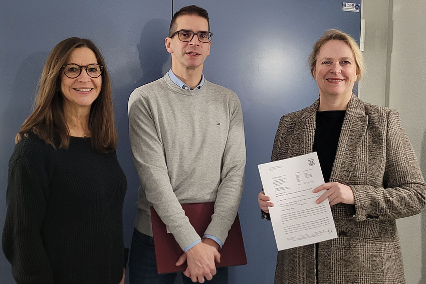 Zwei Frauen und ein Mann stehen vor einer geschlossenen Tür. Die rechts stehende Frau hält ein Schriftstück in der Hand, der Mann links daneben eine Aktenmappe.